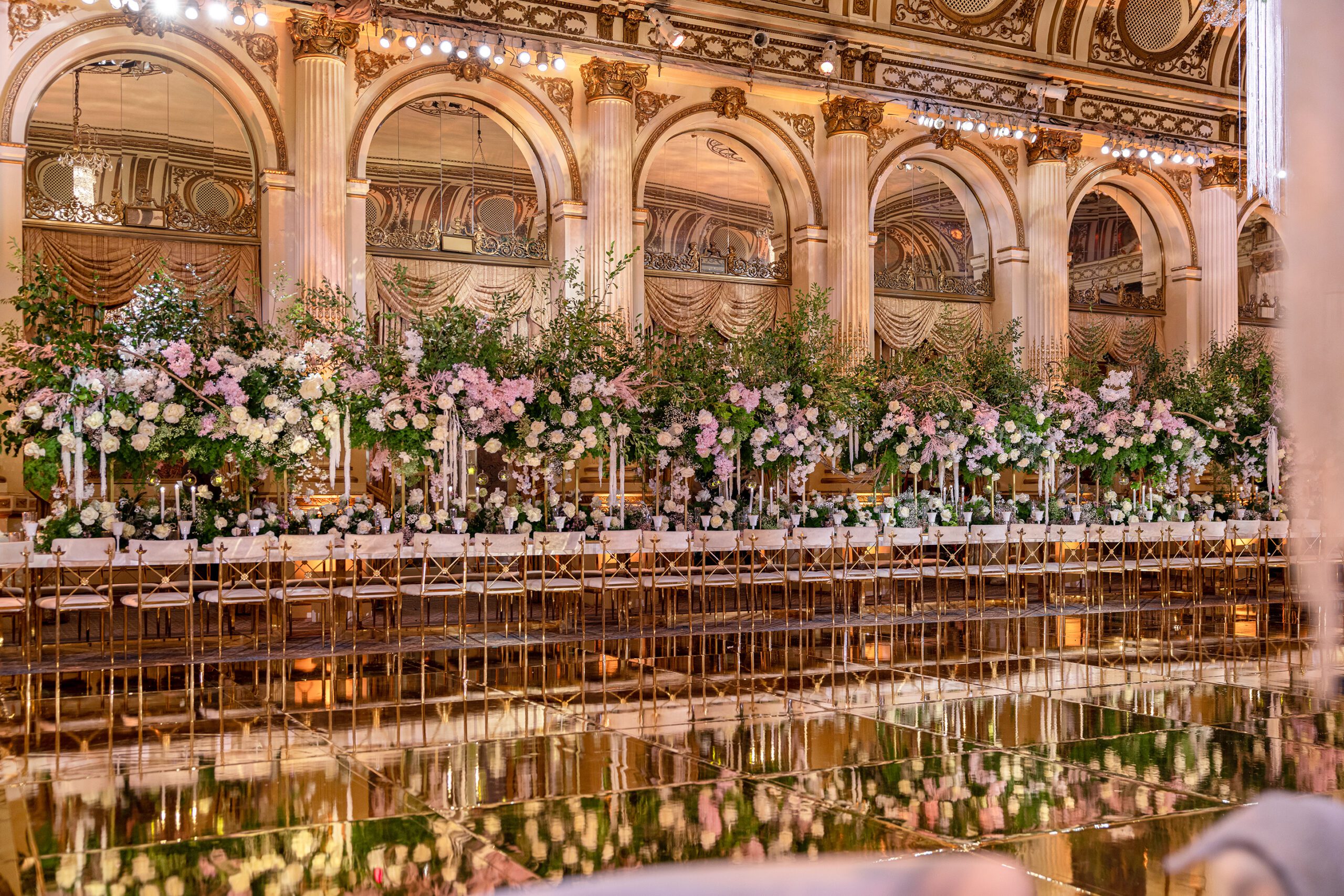 Elegant wedding reception hall adorned with long tables set with white linens, surrounded by abundant floral arrangements crafted by a luxury wedding florist, reflective gold floors, and ornate architectural details.
