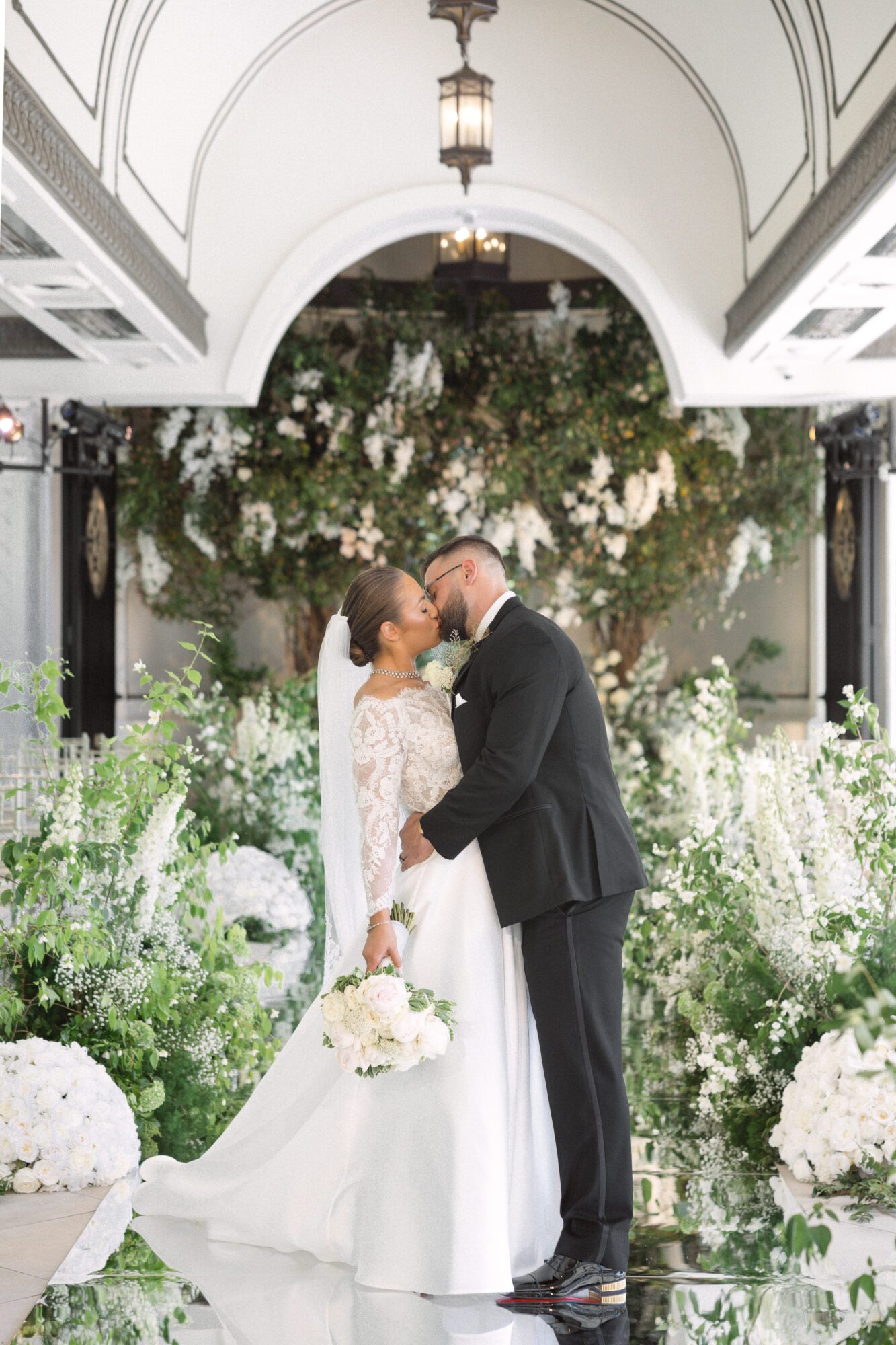 A bride and groom share a kiss amidst a lavish floral setting with white and green blooms designed by a luxury wedding florist in an elegant indoor venue with arched white ceilings.
