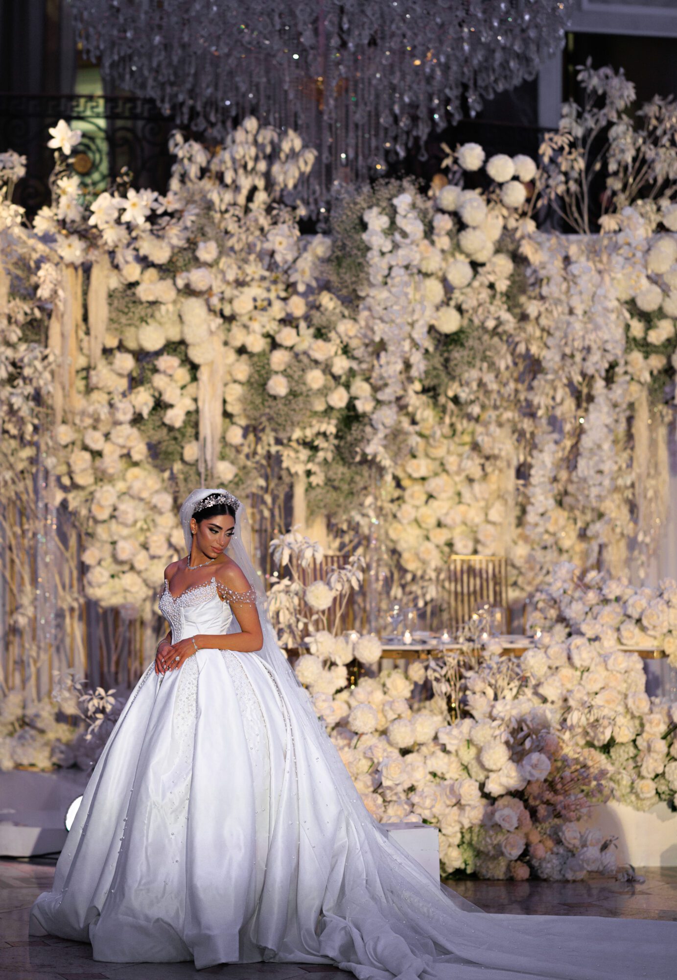 A bride stands amid a luxurious floral backdrop created by a luxury wedding florist, wearing an elegant white gown with a sweeping skirt and pearl accessories, her hair styled in an intricate updo.