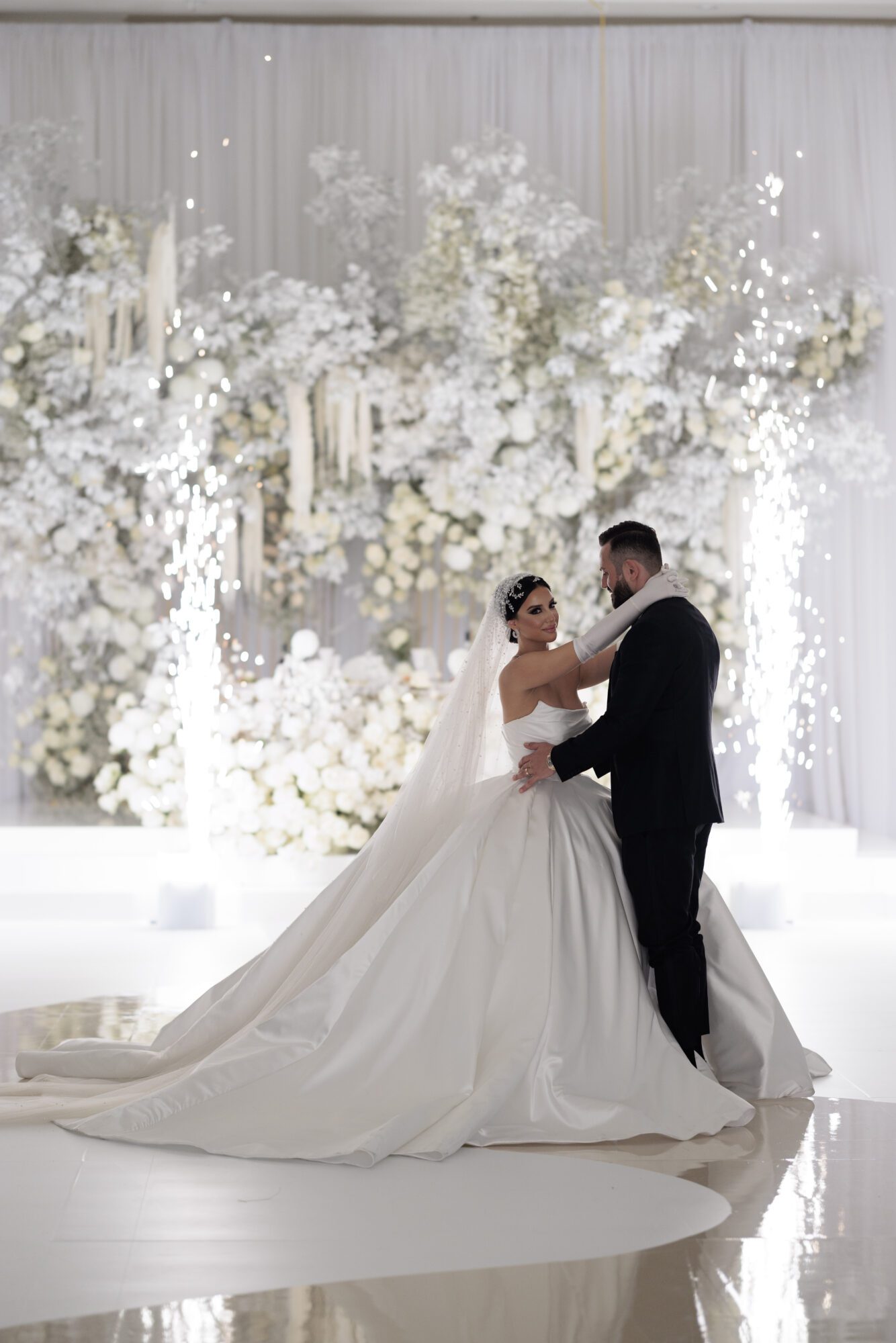 A bride in a flowing white gown and a groom in a black suit share a tender moment in a lavish wedding hall adorned with luxury white flowers and twinkling lights.
