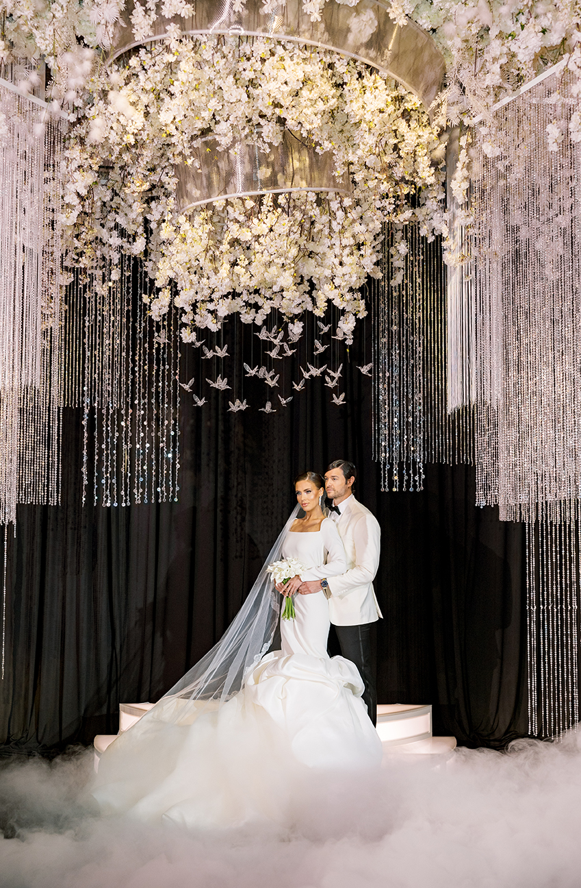 Photo of bride and groom on a stage with a large ceiling display of white florals with crystals hanging from the ceiling including a black draped background and fog throughout the floor