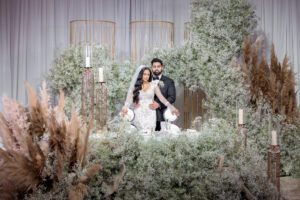 A newlywed couple standing behind a lavishly decorated table with floral arrangements and candles, surrounded by tall pampas grass and white blooms.