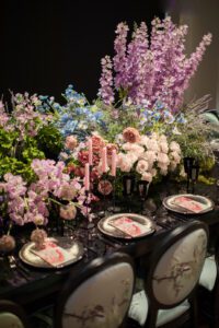 A lavish table setting featuring ornate floral arrangements with purple, pink, and white flowers. The table is set with elegant plates, black stemware, and pink candles in black holders. The backdrop is dark, highlighting the vibrant colors and intricate details.
