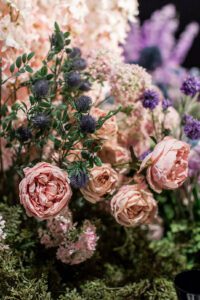 A vibrant floral arrangement featuring pink peonies and purple thistles, surrounded by lush greenery and other assorted blossoms. The flowers are densely packed, creating a rich tapestry of colors and textures.