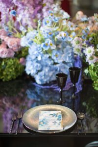 A luxuriously decorated dining table with a glittery gold plate, black cutlery, black glassware, and a floral-themed menu card. In the background, there is a large arrangement of blue and purple flowers, including blue hydrangeas and other assorted blossoms.