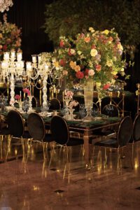 A lavishly set dining table with black and gold chairs, elegant crystal candelabras, and a large, colorful floral arrangement in a tall vase, creating an opulent and festive atmosphere.