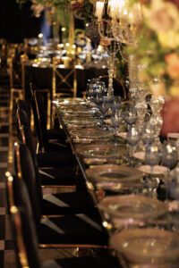 A long, elegant dining table is set with glass plates, blue-tinted goblets, and crystal candelabras. Black and gold chairs line the table, and floral arrangements with candles decorate the center. The setting is dimly lit.