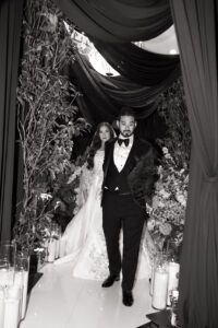 A bride in a white gown and a groom in a tuxedo walk together down an aisle adorned with flowers, greenery, and draped fabric, surrounded by candles. The image is in black and white.