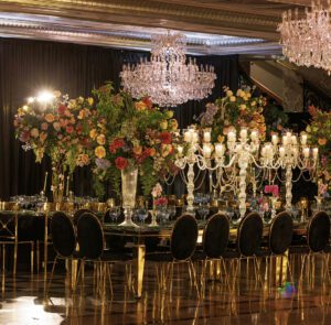 A lavishly decorated banquet table features tall floral arrangements, crystal candelabras, and black velvet chairs under sparkling chandeliers in an elegant, formal setting.