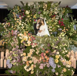 A bride and groom embrace and kiss in front of a lush, colorful wall of flowers, surrounded by abundant floral arrangements and rows of lit candles.