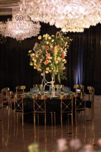 A round table with gold chairs set for a formal event, featuring an elaborate floral centerpiece with colorful flowers. Large crystal chandeliers hang above, and dark curtains provide an elegant backdrop.
