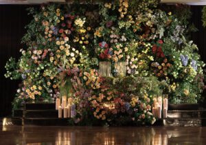 A lush, vibrant floral wall filled with various colorful flowers and greenery serves as a backdrop. In front, numerous lit pillar candles create a soft, romantic glow, with two ornate chairs placed at the center.