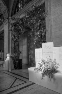 A black and white photo of an elegant wedding venue entrance featuring a floral arch, a large sign with "The Perfettos, May 13, 2023," and a nearby table holding a tiered wedding cake.