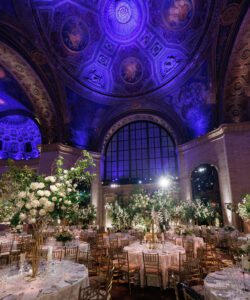 Elegant banquet hall with ornate, domed ceiling illuminated in purple light. Tables are set with white linens, gold chairs, and tall floral centerpieces, creating a luxurious, formal atmosphere. Large arched windows in the background.