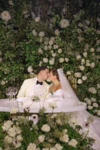 A bride and groom kiss at a round table decorated with white flowers and glassware, surrounded by lush greenery and white floral arrangements, both dressed in formal wedding attire.