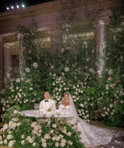 A bride and groom sit at a white table decorated with white flowers and lush greenery, surrounded by grand columns and abundant floral arrangements; the bride wears a long white gown and veil.