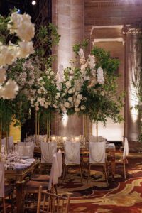 Elegant dining area with gold tables and chairs, white draped chair covers, and lavish floral centerpieces featuring white flowers and greenery, set in a grand, columned room with ornate decor.