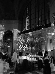 Black and white photo of an elegant indoor event with tables, chairs, and tall flower arrangements. Large windows reflect city skyscrapers at night; people are walking and mingling in formal attire.