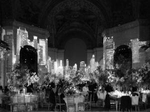A black-and-white photo of an elegant event in a grand hall, with guests seated at decorated tables surrounded by flowers; city skyscrapers are projected onto the walls, creating a dramatic urban backdrop.