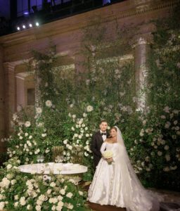 A bride and groom stand together in formal wedding attire in front of a lush, green floral backdrop, with a decorated table holding candles and glasses in the foreground. The scene is elegant and romantic.