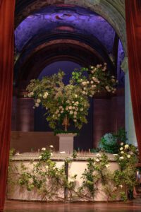 An ornate indoor scene features a large floral arrangement with white flowers and greenery on a pedestal behind a bar decorated with more flowers, set beneath an arched ceiling with dramatic lighting.
