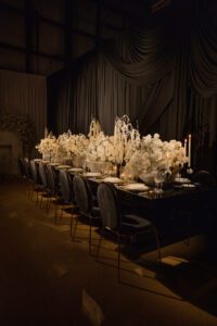 A long, elegant dining table set for a formal event features dark chairs, white floral centerpieces, candles, and dramatic draped black and gray fabric in the background, creating a luxurious and moody atmosphere.