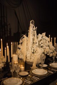 An elegant table setting with white plates, gold-rimmed glasses, tall lit candles, and a lush centerpiece of white flowers and cascading cream-colored foliage, set against a dark, draped background.