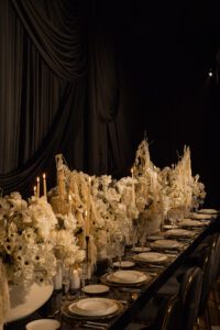 A luxurious dining table set with elegant white plates, gold-trimmed glassware, tall candles, and abundant white floral arrangements, set against lavish black drapes in a dimly lit room.