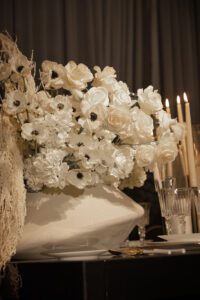 A large arrangement of white flowers, including roses and anemones, sits on a table decorated with crystal glassware, gold utensils, and lit taper candles against a dark, draped background.