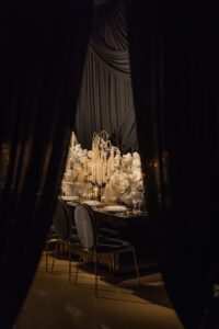A luxurious dining table with elegant black chairs, white floral arrangements, and candles is framed by dark, draped curtains, creating an intimate and sophisticated atmosphere.