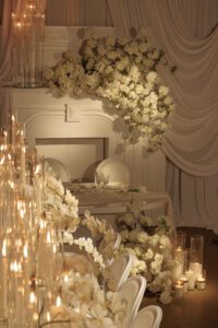 Elegant wedding table setting with white flowers, candles in glass holders, and draped white fabric. Soft lighting creates a romantic atmosphere, with floral arrangements cascading over the table and surrounding a fireplace.