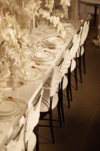 Elegant table setting with white plates and gold-rimmed glassware, white floral arrangements, and white chairs with wing-shaped backs, arranged neatly along a long, glossy white table.