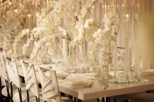 Elegant table setting with white plates, crystal glasses, and tall glass vases filled with cascading white orchids, creating a luxurious and sophisticated atmosphere. White chairs and soft lighting enhance the scene.