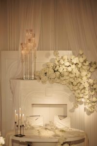 A romantic wedding table setting featuring two chairs, a round table with white decor, tall glass candle holders, and abundant white flowers cascading over a white backdrop with draped fabric.