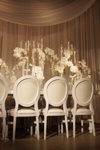 An elegant table setting with white chairs, tall glass vases holding white orchids, and draped white fabric in the background. Warm lighting creates a soft, sophisticated atmosphere.