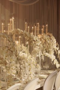 A luxurious table setting featuring cascading white orchids, tall glass candle holders with lit candles, elegant glassware, and white plates, set against soft beige draped curtains.