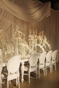 An elegant dining table with white chairs, draped white fabric in the background, tall glass candle holders, and white orchid floral arrangements create a luxurious, monochromatic setting.