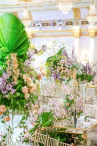 A lavish ballroom decorated with tall, lush floral arrangements featuring pastel flowers and large green leaves, elegant gold chairs, chandeliers, and ornate architectural details in the background.