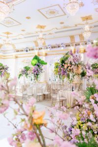 Elegant ballroom decorated with tall floral centerpieces, pastel flowers, lush greenery, gold accents, and chandeliers. White chairs and tables create a luxurious, airy atmosphere for a formal event or wedding reception.