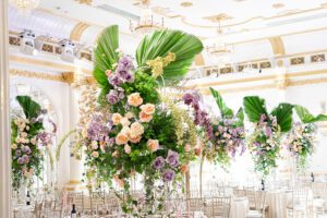 Elegant ballroom decorated with tall floral arrangements featuring purple and peach flowers, lush greenery, and large palm leaves. Crystal chandeliers hang from the ornate, gold-accented ceiling above white tables and chairs.