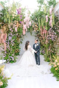 A bride in a long white gown and a groom in a tuxedo stand facing each other under a lush floral arch with pink, purple, and green flowers and greenery, creating a romantic indoor wedding scene.