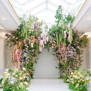 A lush floral archway with cascading green foliage and pastel flowers, including pink, purple, and peach blooms, stands under a glass ceiling in a bright, modern indoor space.