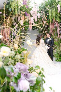 A bride and groom stand facing each other amid lush, colorful flowers and greenery, creating a romantic, whimsical wedding scene. The bride wears a long white gown; the groom is in a black tuxedo.