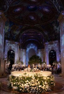 A grand, circular banquet table with abundant white flowers sits under a domed ceiling with ornate, painted artwork in an opulent hall, with guests seated around and elegant lighting illuminating the scene.