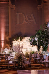 An elegant wedding reception hall features floral centerpieces, crystal chandeliers, and a large "D&A" monogram on the wall. Clear chairs and lush greenery add to the sophisticated atmosphere.