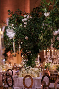 A lavish wedding reception table features a tall, lush centerpiece with greenery and white flowers. Elegant round chairs and tables with white linens and floral arrangements surround the centerpiece. Warm lighting enhances the scene.