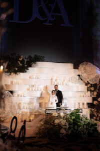 A bride and groom stand together in front of a modern white wedding backdrop decorated with candles, white flowers, and greenery, with large initials "D & A" displayed above them.
