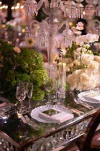 Elegant table setting with crystal glassware, plates, white napkins, lush green and white floral arrangements, and a tall, ornate crystal candelabra centerpiece, creating a luxurious, festive atmosphere.