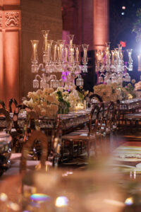 Elegant banquet table set for an event, adorned with crystal candelabras, white floral arrangements, glassware, and mirrored surfaces under warm, ambient lighting, creating a luxurious and festive atmosphere.