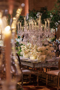 Elegant banquet table decorated with large crystal candelabras, lush white flower arrangements, and reflective surfaces, surrounded by chairs in a lavish, warmly lit venue.